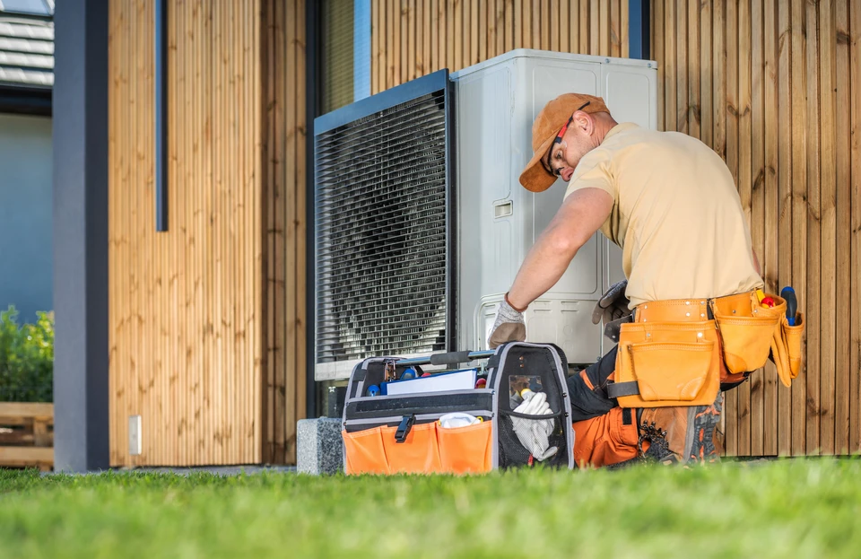 Technicien en climatisation travaillant sur une unité extérieure de pompe à chaleur devant un bâtiment moderne à bardage bois