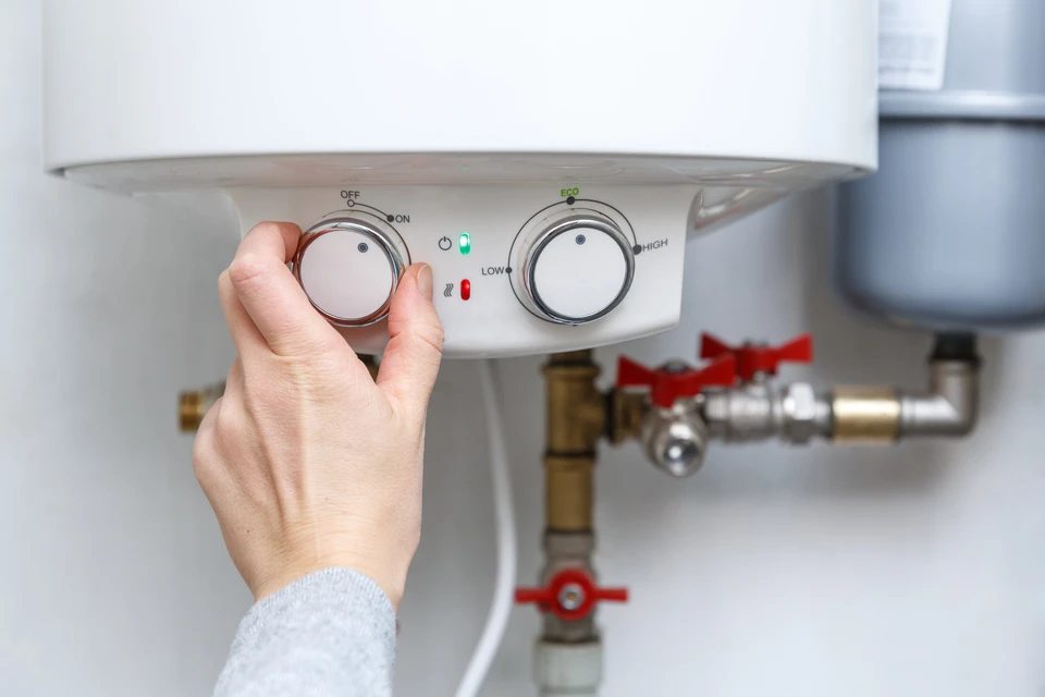 Une main ajuste le thermostat d'un chauffe-eau domestique blanc fixé au mur